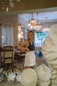 Blonde woman in stylish attire stands in a decorated room with chandeliers.
