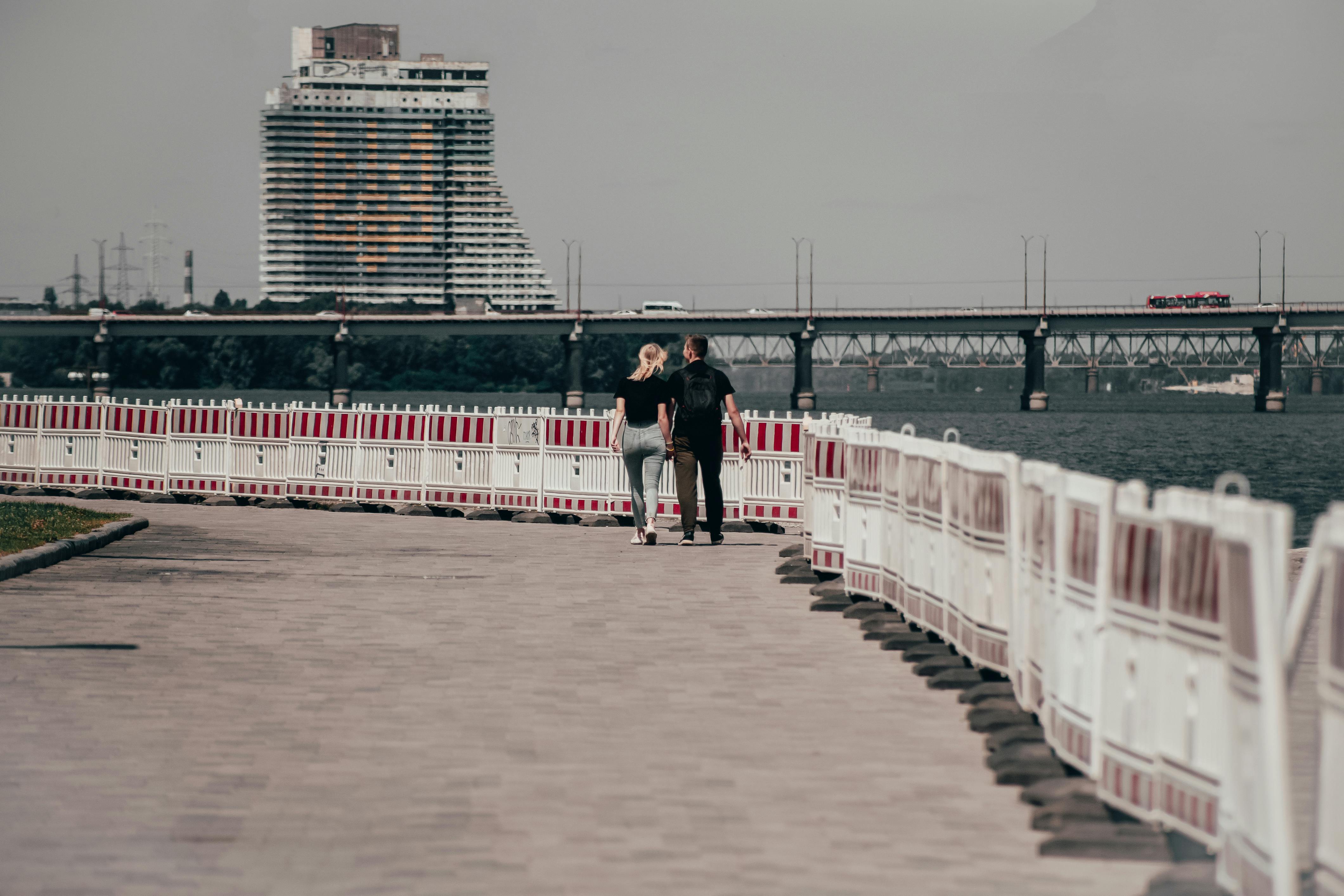 A couple walks along a riverside promenade with a bridge and modern building in the background