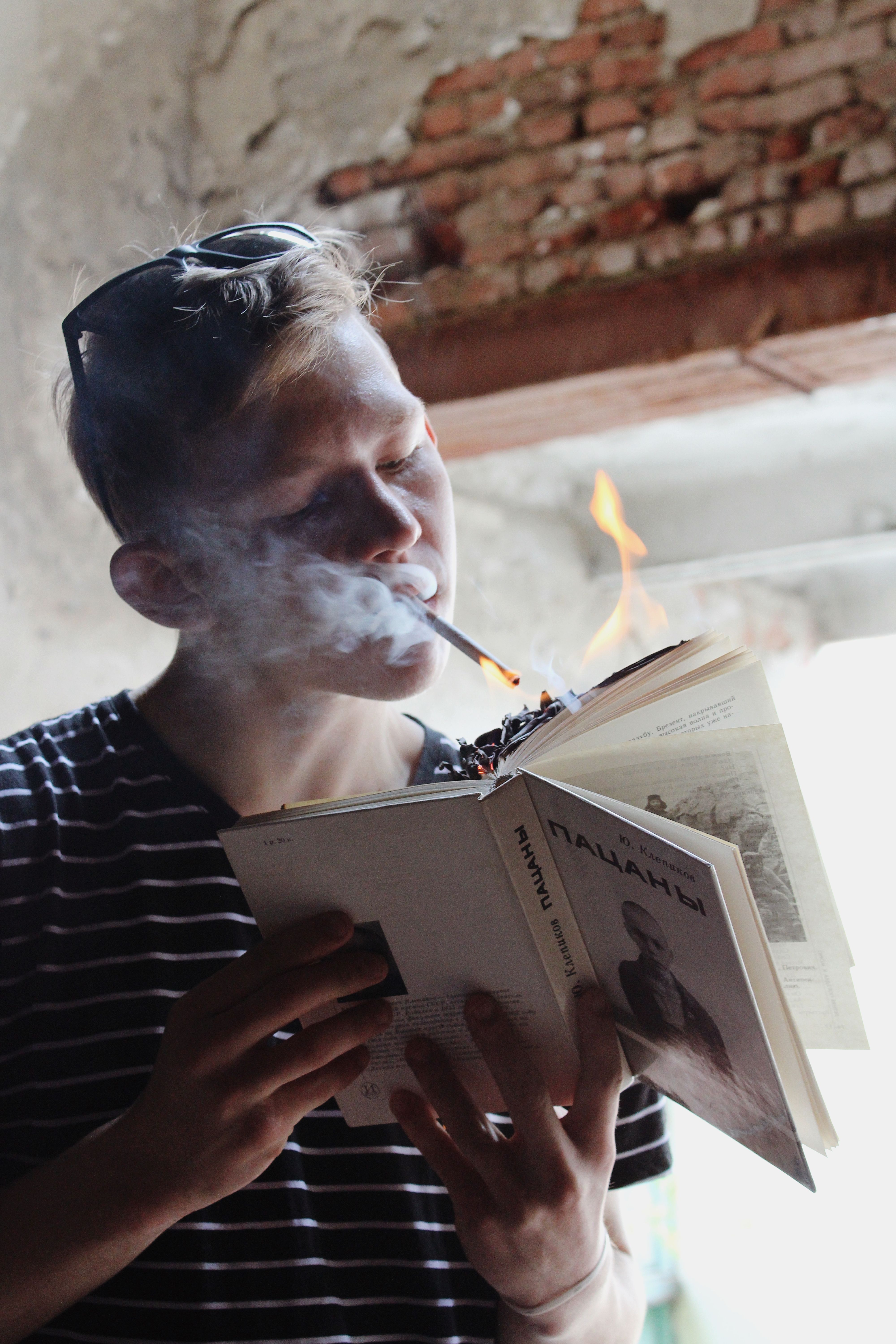 A young man smokes a cigarette while a book burns indoors, creating a dramatic scene.