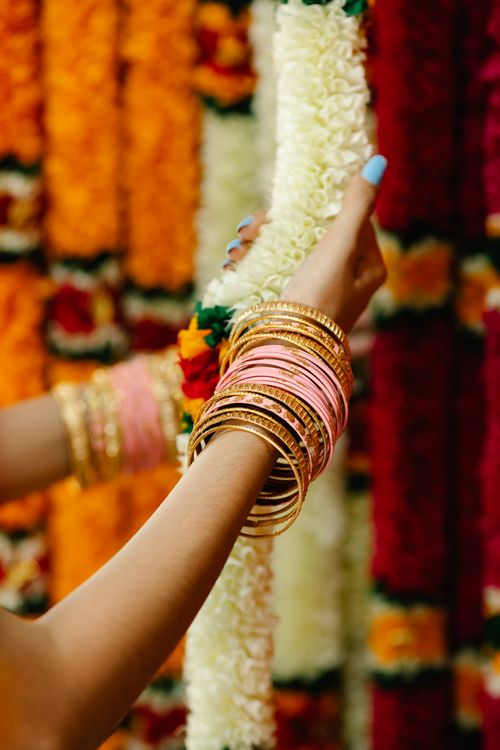 Free Colorful display of a woman's hand with bangles holding a vibrant flower garland against a floral backdrop. Stock Photo