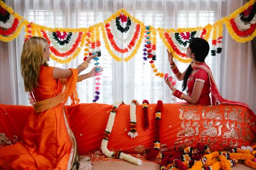 Two women in traditional attire decorating a room with colorful floral garlands.