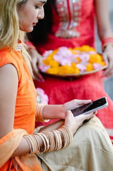A woman in vibrant traditional attire uses a smartphone while another holds a tray of flowers.