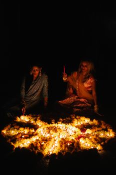 Two women celebrating Diwali at night with candles and traditional attire, creating a rangoli.