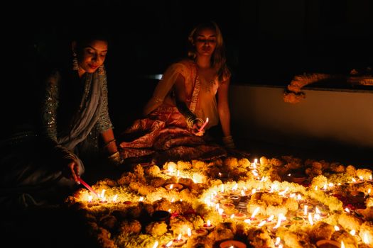 Two women in traditional clothing light candles for Diwali amidst flower arrangements.