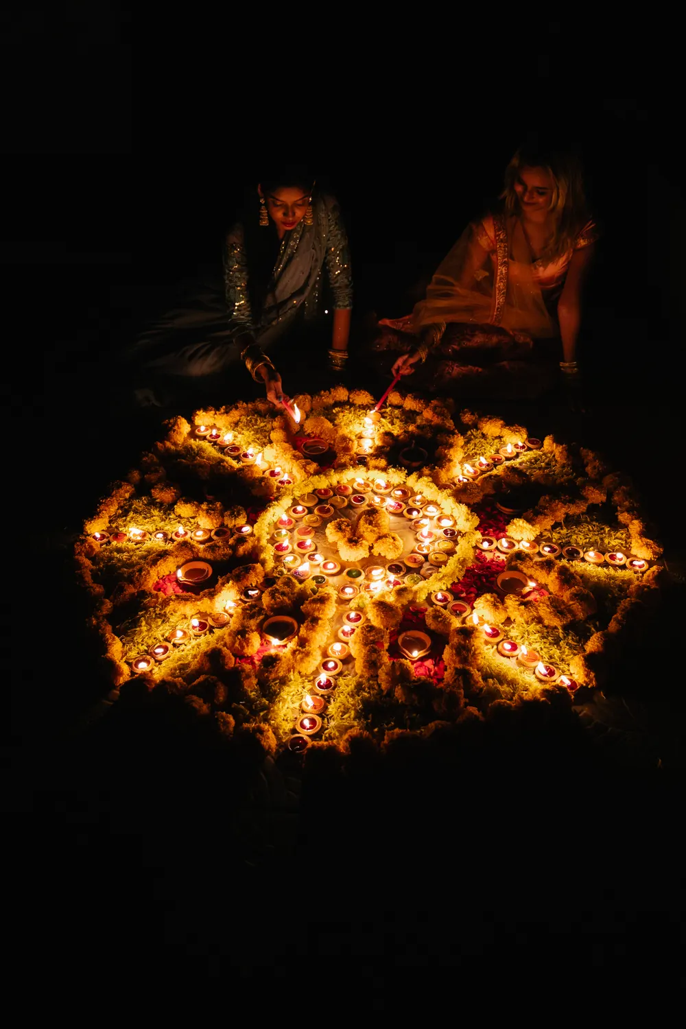 Two women creating a vibrant rangoli design surrounded by candles