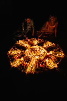 Two women in traditional attire create a vibrant Rangoli surrounded by candles for Diwali celebration.