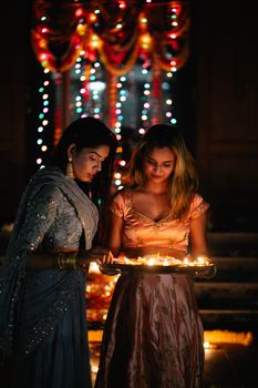 Two women in traditional attire holding lighted diyas at a night ceremony.