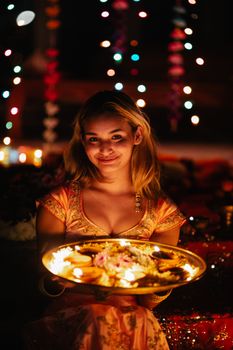 A young woman holds a decorated plate with candles during an Indian festival at night.