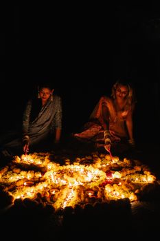 Two women in traditional attire celebrate Diwali by decorating rangoli with candles.