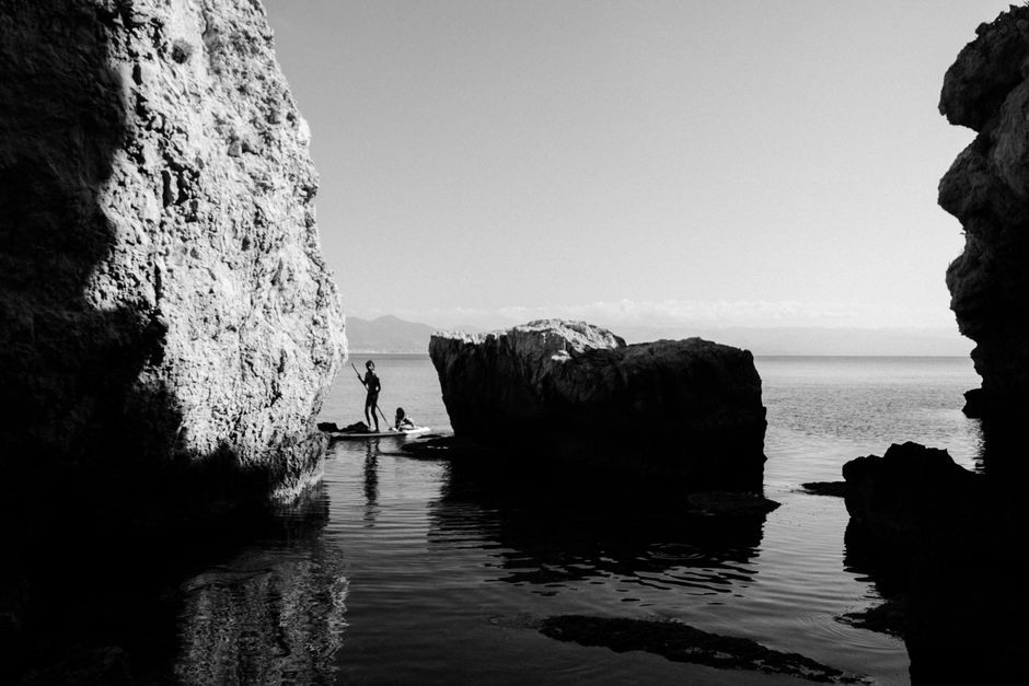 Dramatic black and white photo featuring a paddleboarder between coastal rocks on a calm sea.