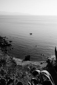 A tranquil black and white photograph of a coastal landscape featuring a solitary boat on calm water.