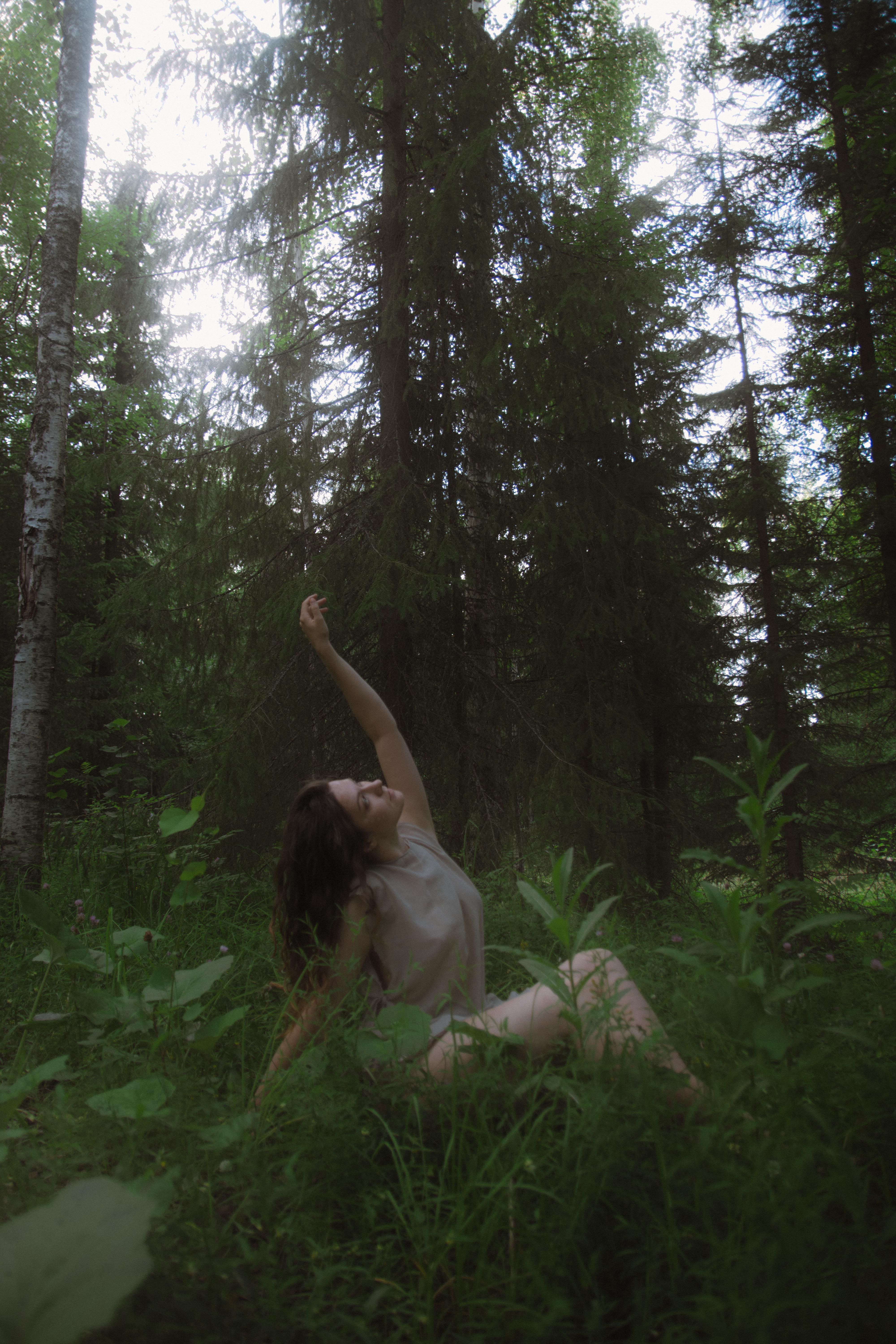 A woman sits gracefully on forest grass reaching towards the sky surrounded by tall trees.