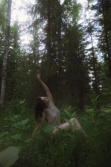 A woman sits gracefully on forest grass reaching towards the sky surrounded by tall trees.