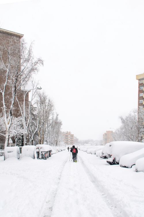 Free Photograph of a snow-covered city street with people walking, lined by buildings and trees. Stock Photo