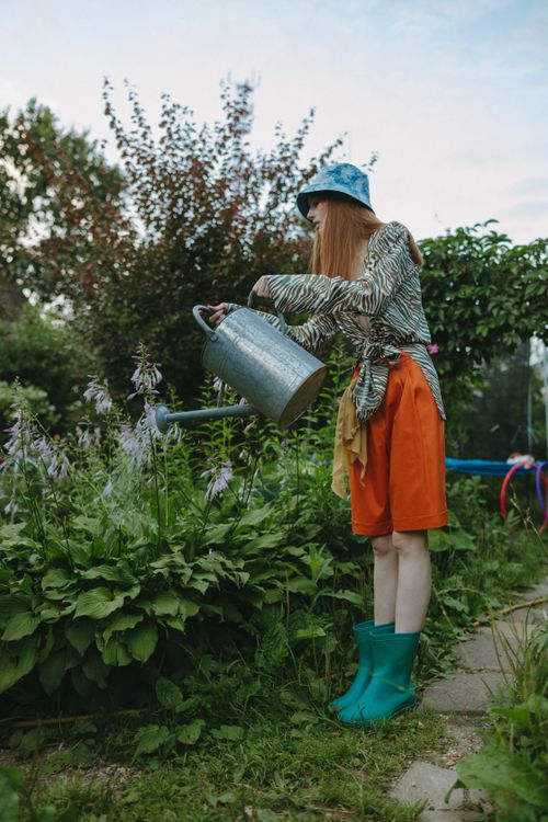 Free A stylish woman in a bucket hat waters plants in a lush garden, showcasing modern gardening fashion. Stock Photo