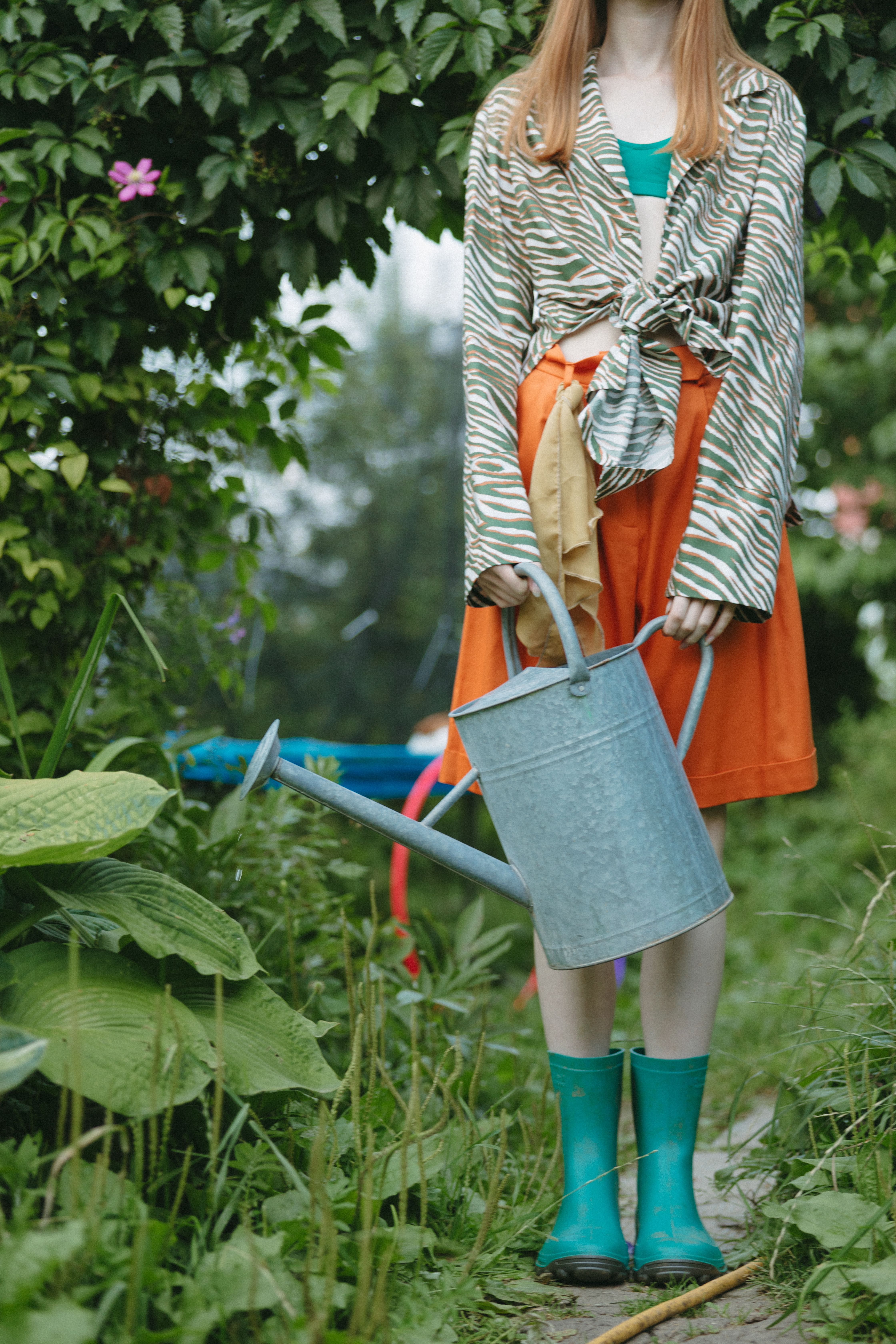 A fashionable woman holding a watering can in a lush garden setting.
