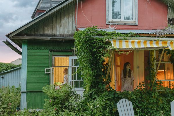 Host standing at the lit window of a green timber cottage at dusk — photo by Yaroslav Shuraev on Pexels