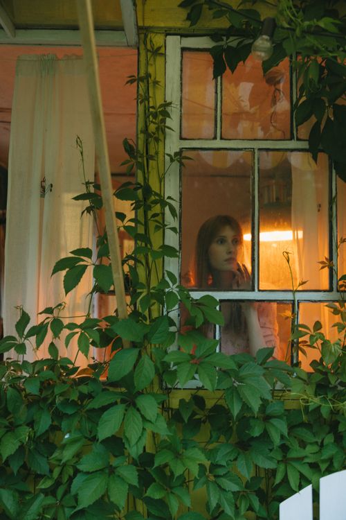 Free A woman looks thoughtfully outside a window framed by lush green plants during a tranquil moment indoors. Stock Photo