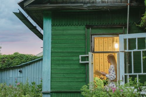 Free A serene scene of a woman reading inside a wooden house at sunset. Stock Photo