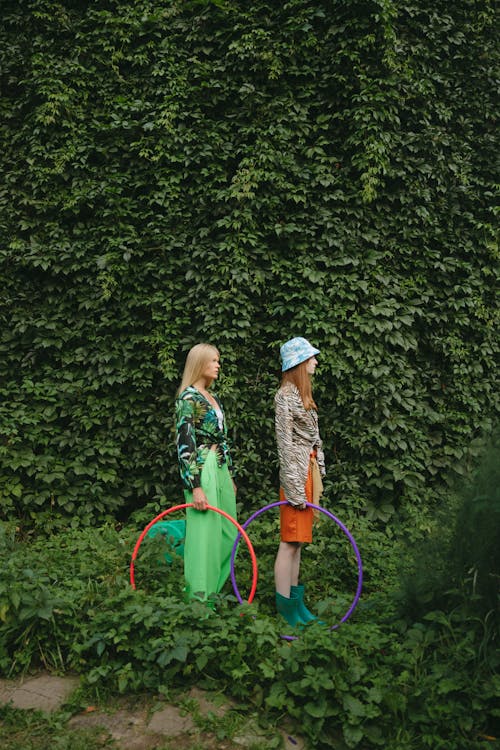 A Side View of Women Standing Near the Green Plants while Holding Hula Hoops