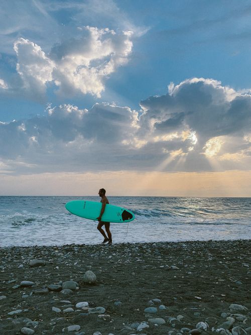 Free A surfer carrying a surfboard along a rocky beach with dramatic sunset clouds and sunbeams over the ocean. Stock Photo
