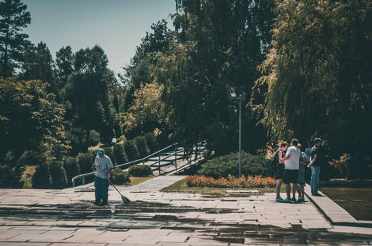 A park setting with a group of people and worker cleaning the walkway.