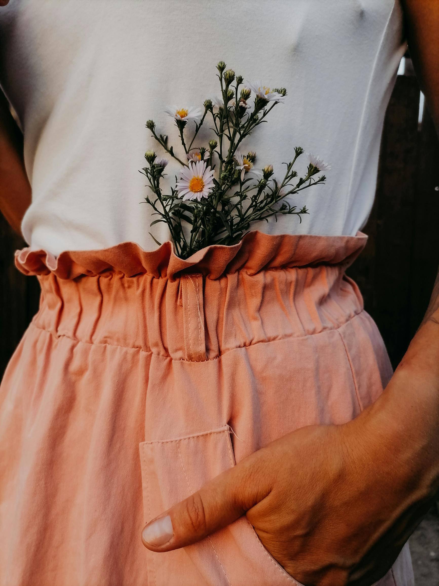 Close-up of a person with daisies tucked into a waistband, hand in pocket, emphasizing a warm and natural style.