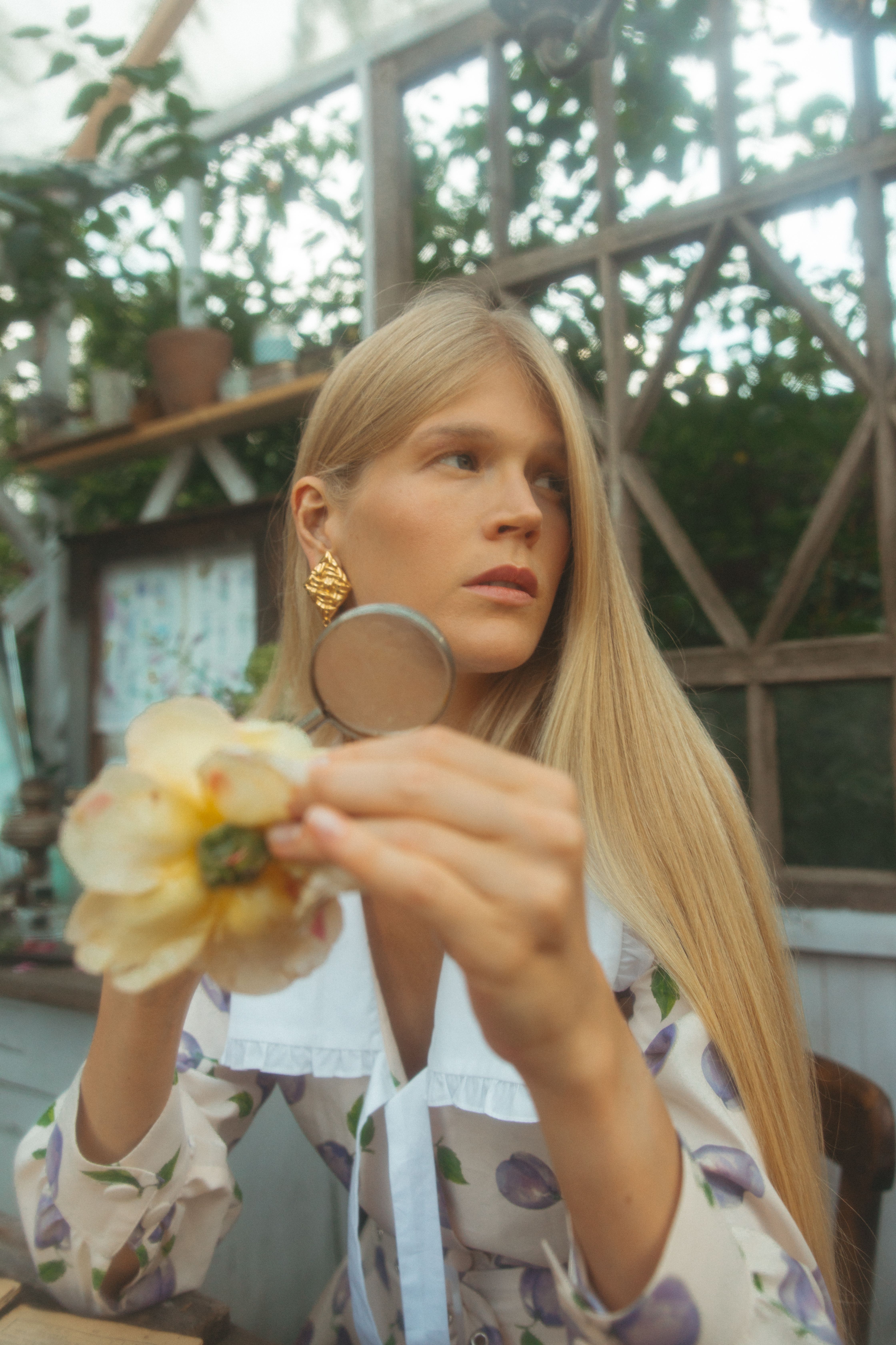 Woman in a floral dress with a magnifying glass inspecting a flower inside a greenhouse.