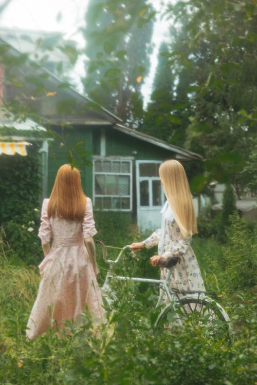 Free Two women in vintage dresses stand beside a bicycle near a country house in a serene countryside setting. Stock Photo