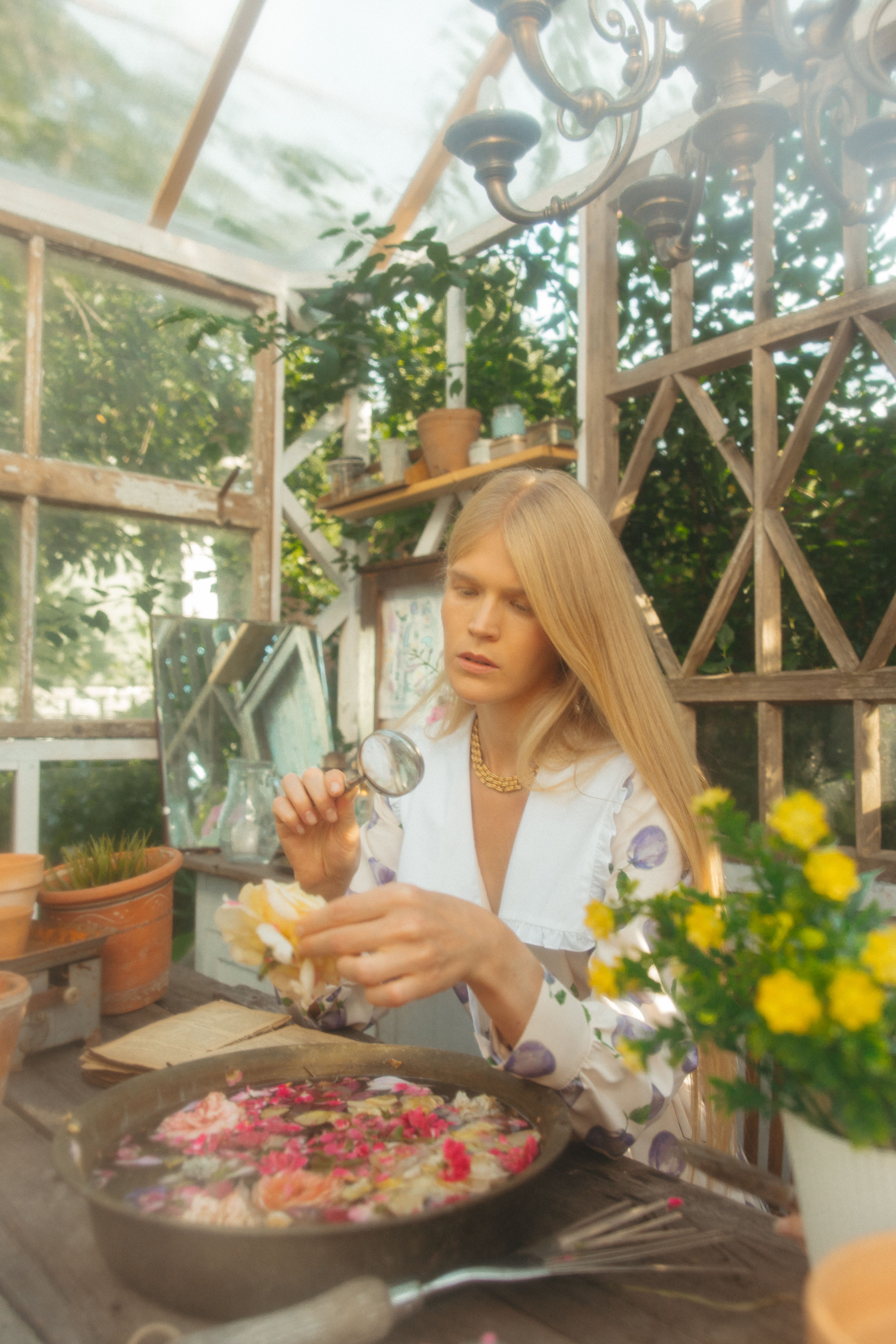 A woman examines flowers with a magnifying glass inside a sunlit greenhouse.
