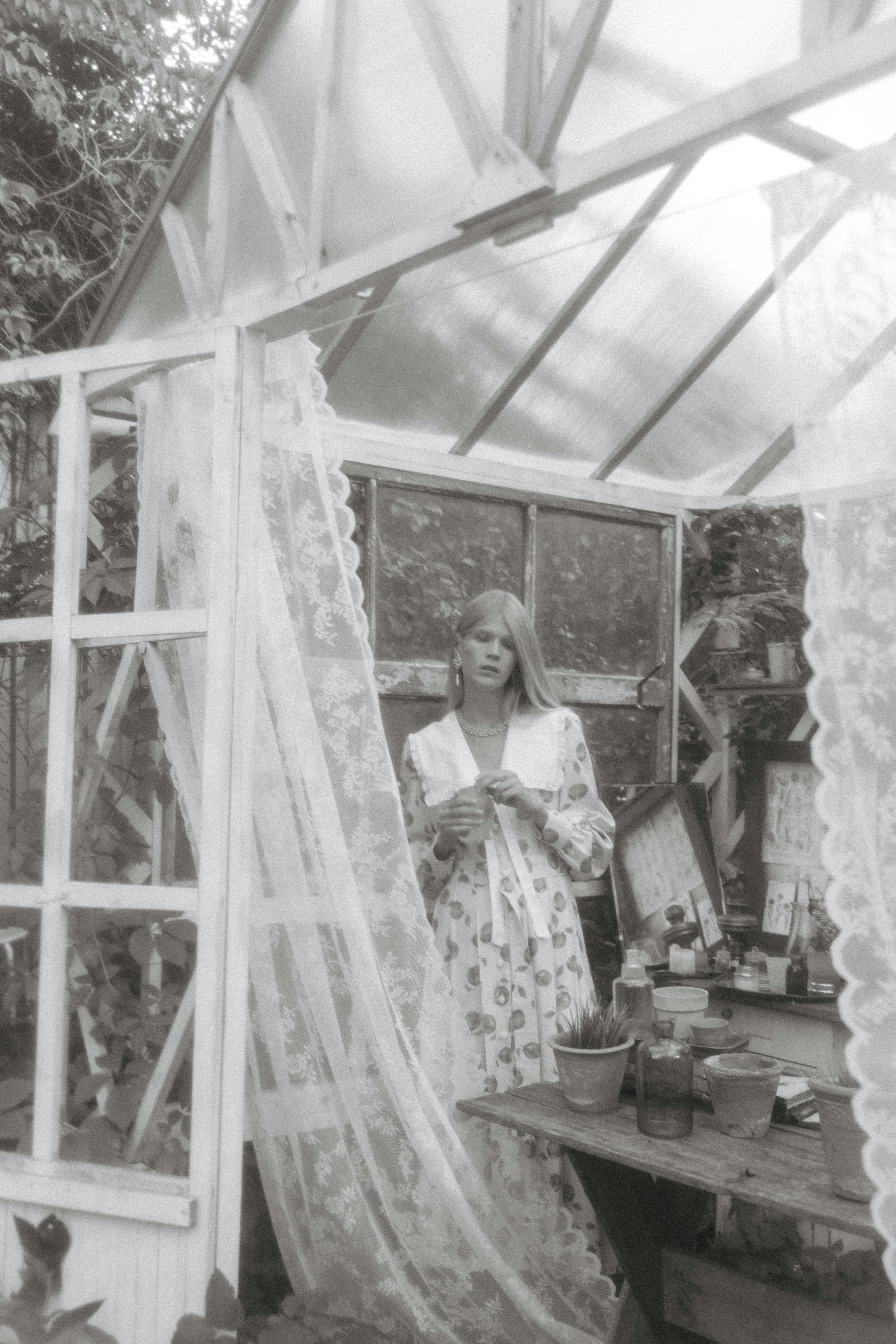 Serene black and white image of a woman in a greenhouse with lace curtains.