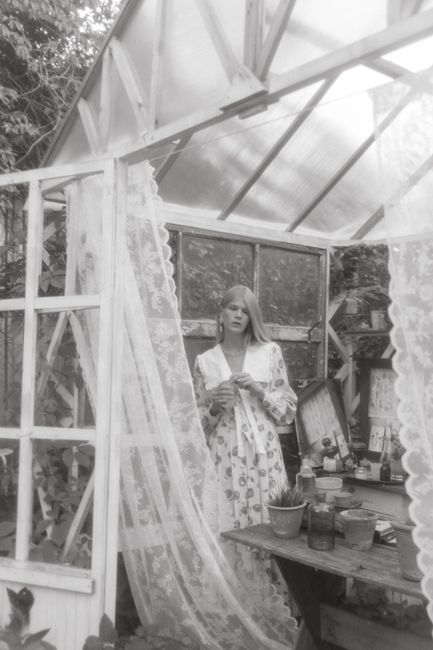 Serene black and white image of a woman in a greenhouse with lace curtains.
