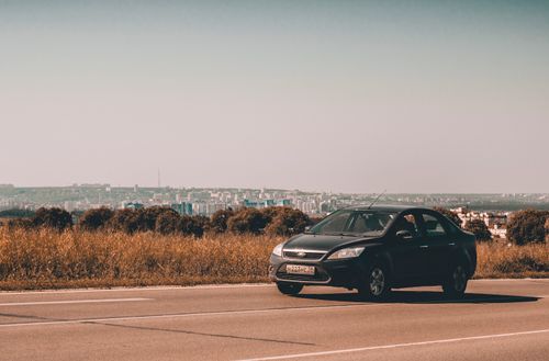 Free A black sedan travels on a country road with a cityscape view in the background under a clear sky. Stock Photo