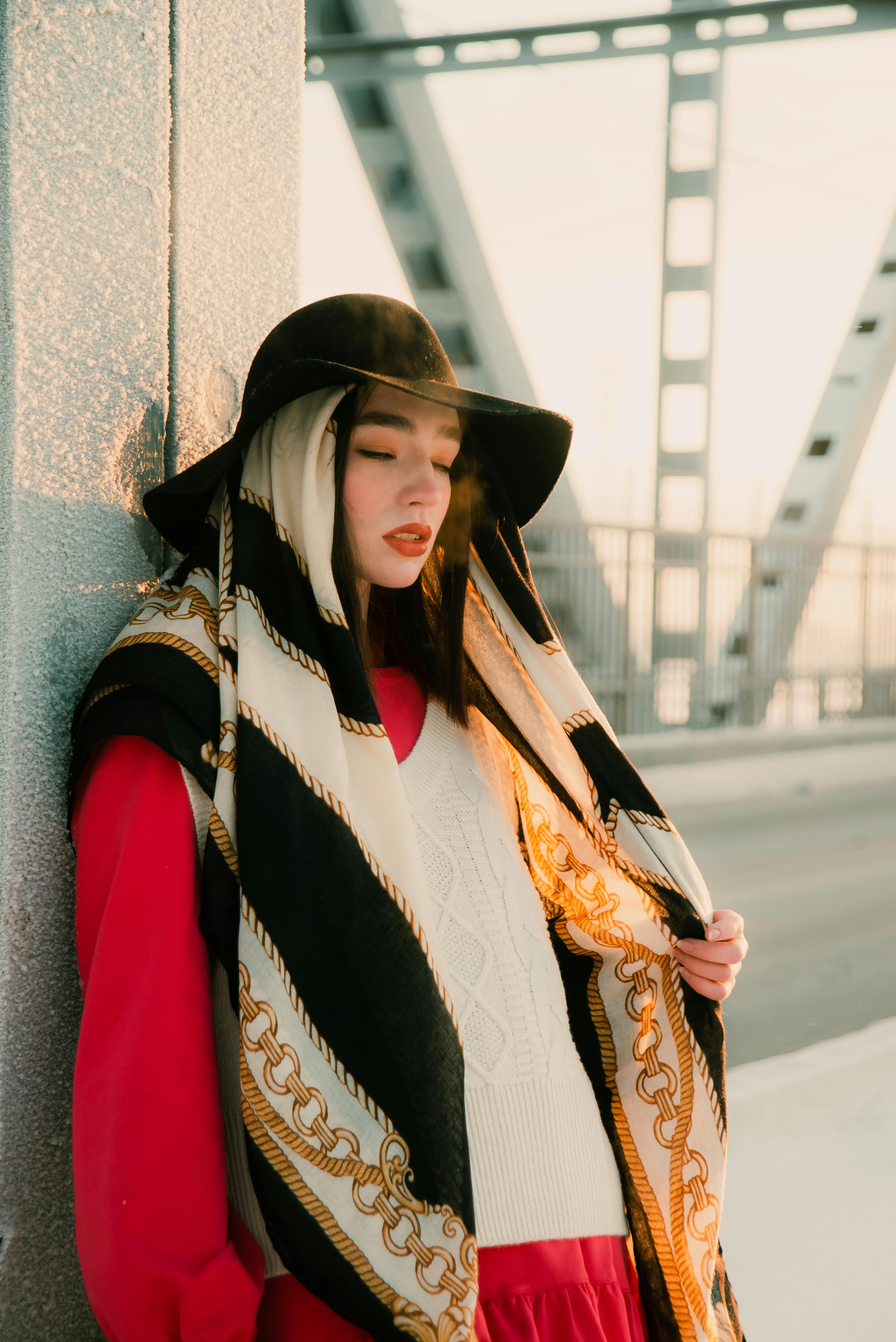 Free Fashionable woman in scarf and hat leaning against bridge with eyes closed, golden light. Stock Photo