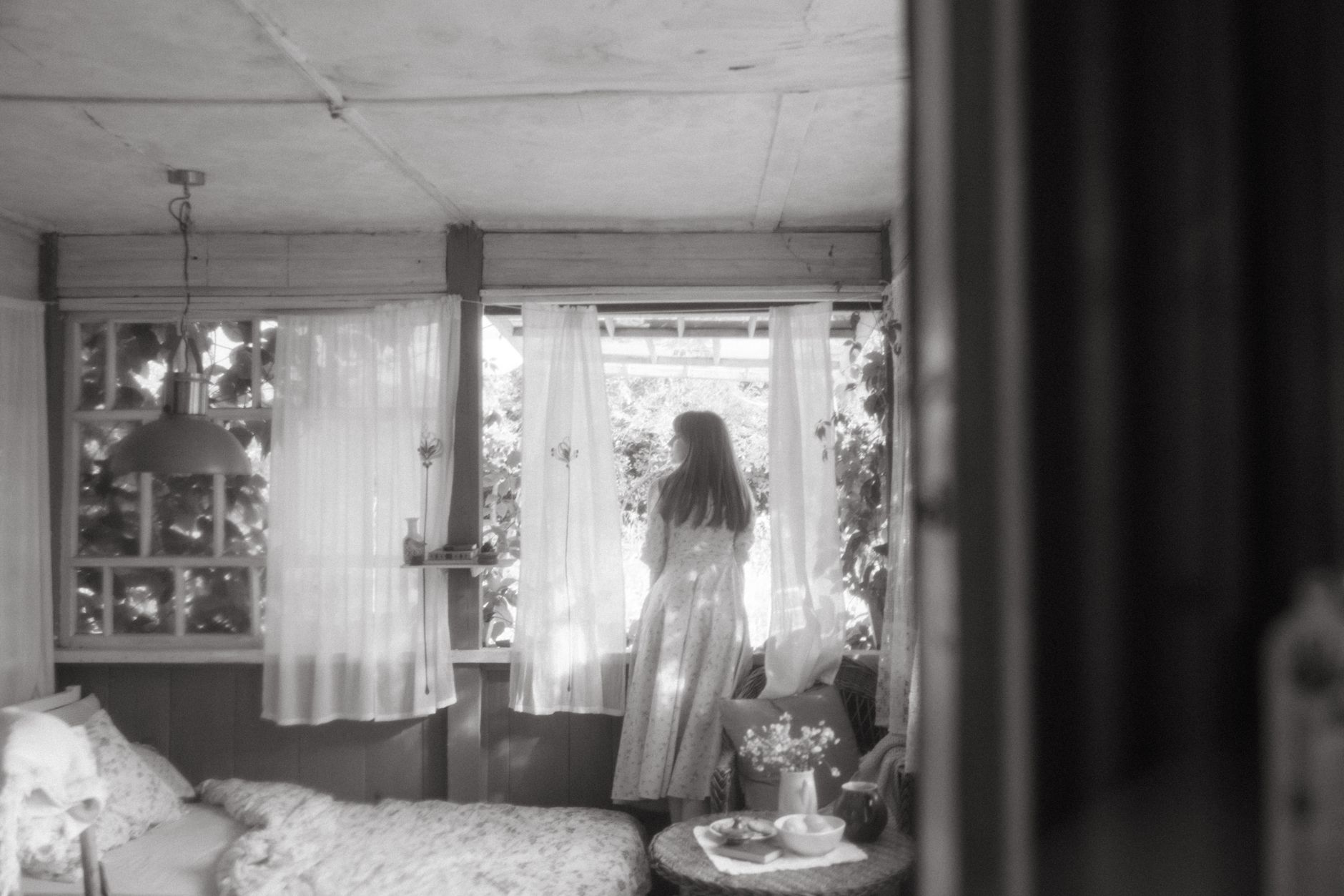 Black and white image of a woman looking out from a cozy, vintage interior.