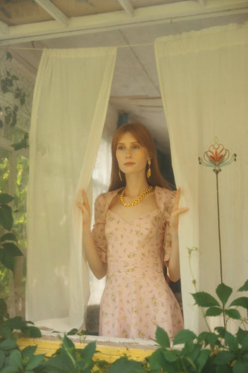 Woman in a White Dress and Gold Necklace Holding the Curtains