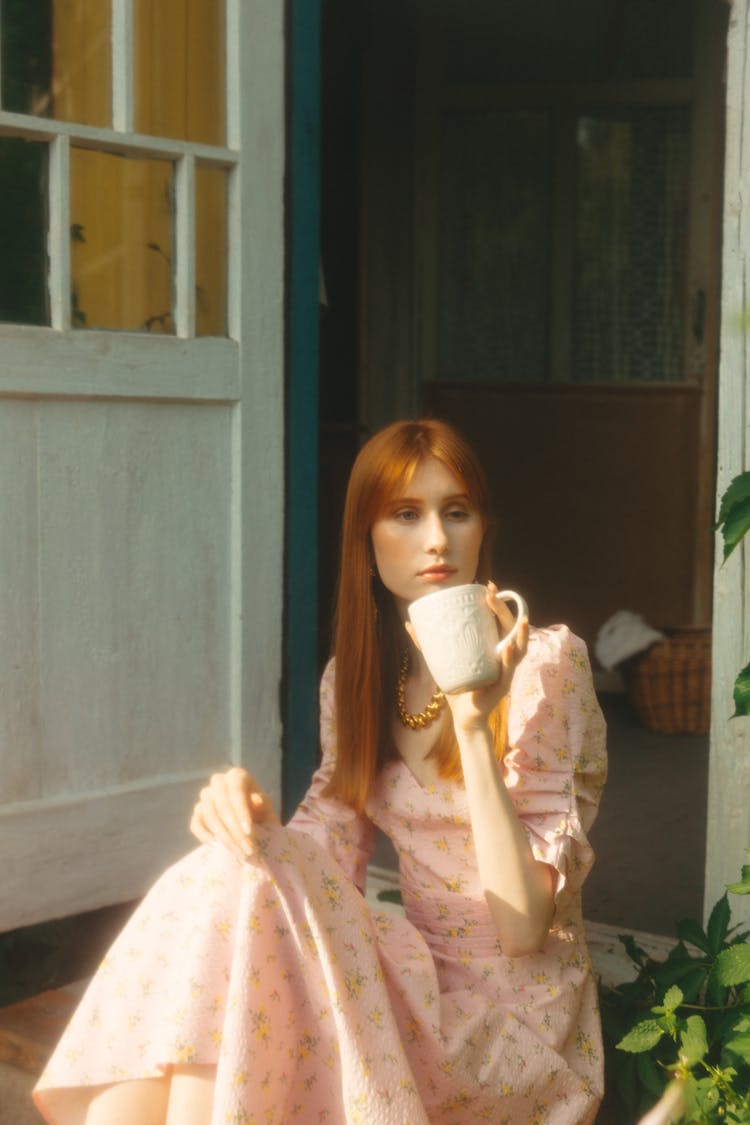 Woman In A Pink Dress Sitting Near The Door Drinking From A Mug