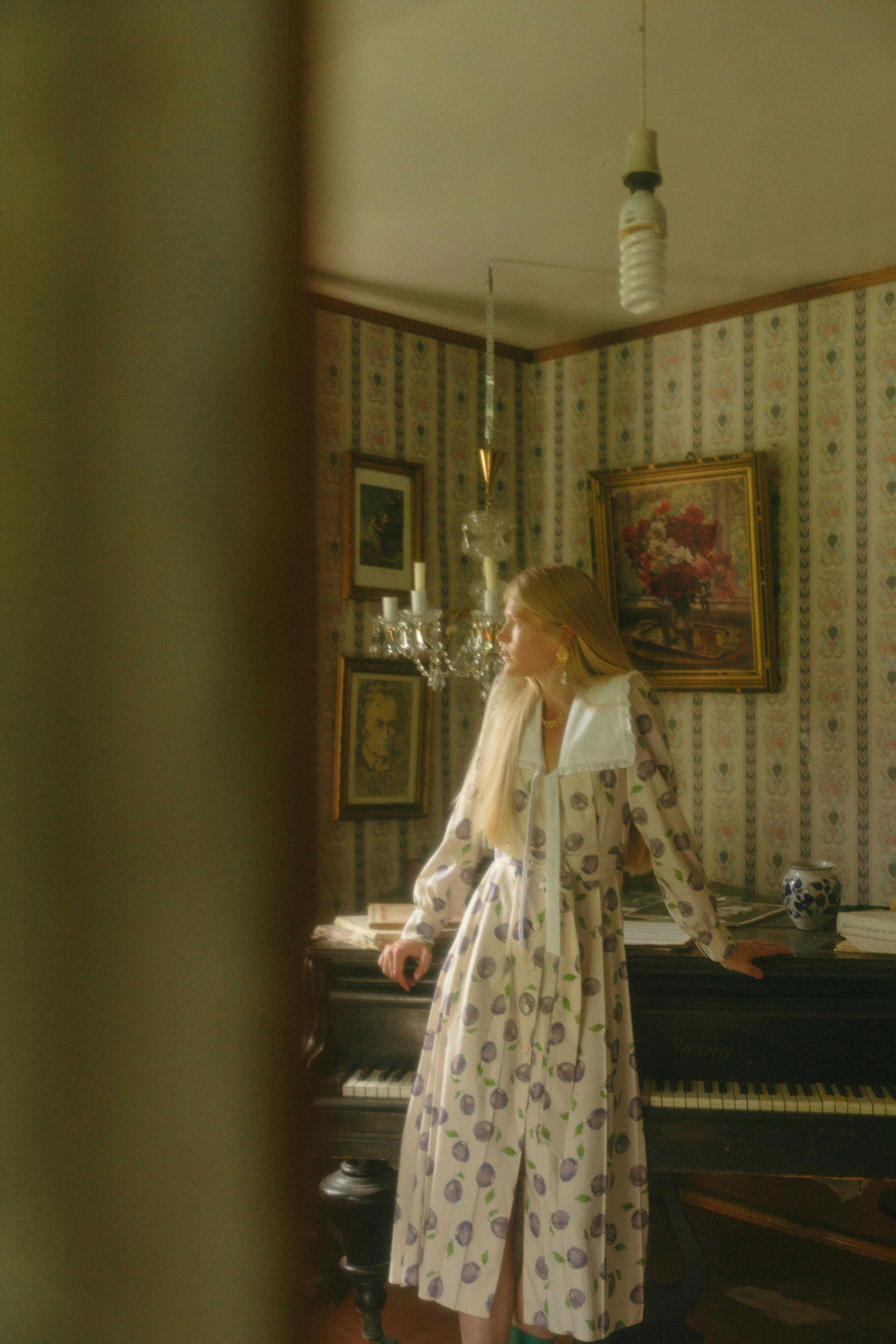 Blonde woman in retro dress leans on piano in a vintage-styled room.