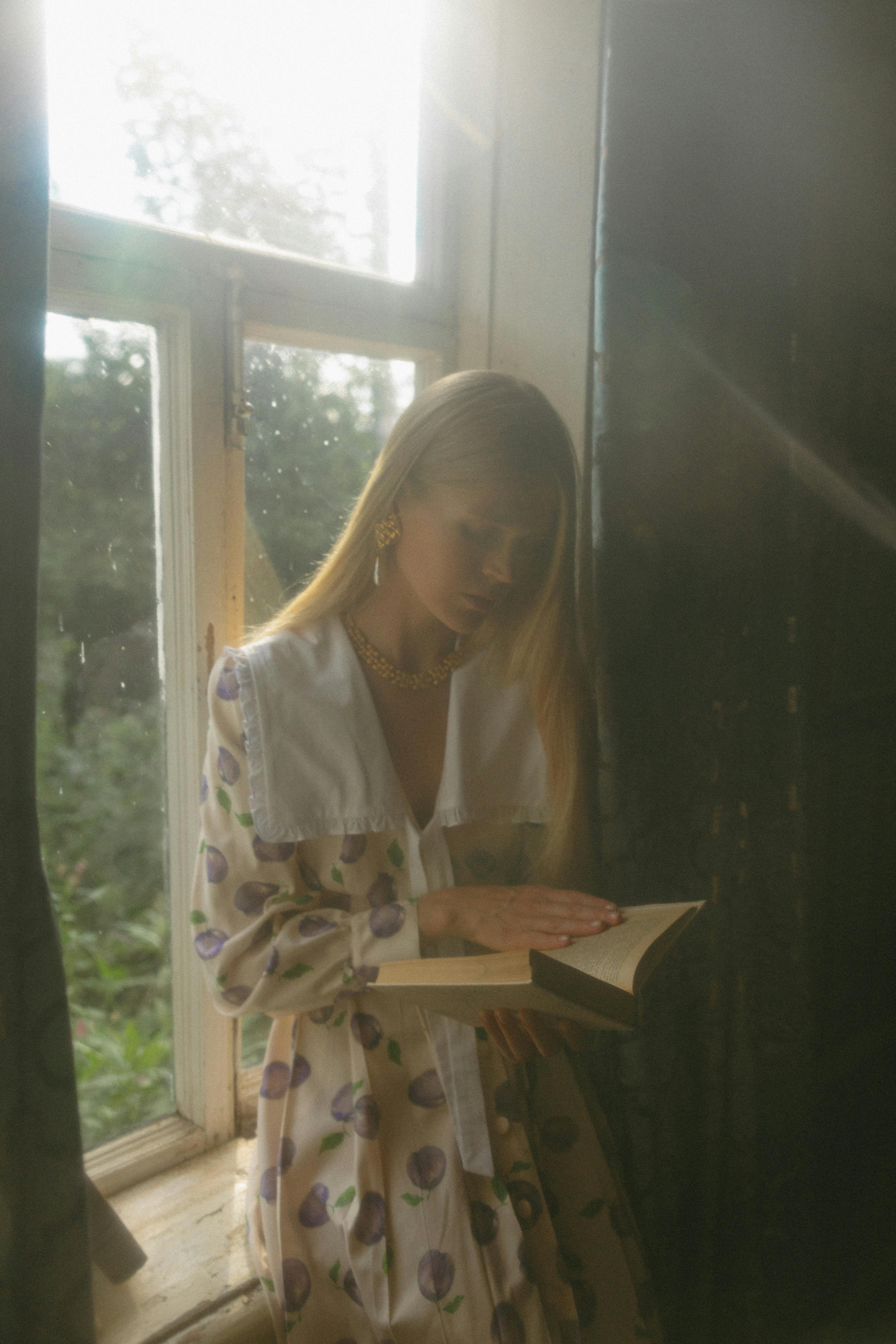 A young woman in stylish attire reads a book by a sunlit window, creating a serene atmosphere.