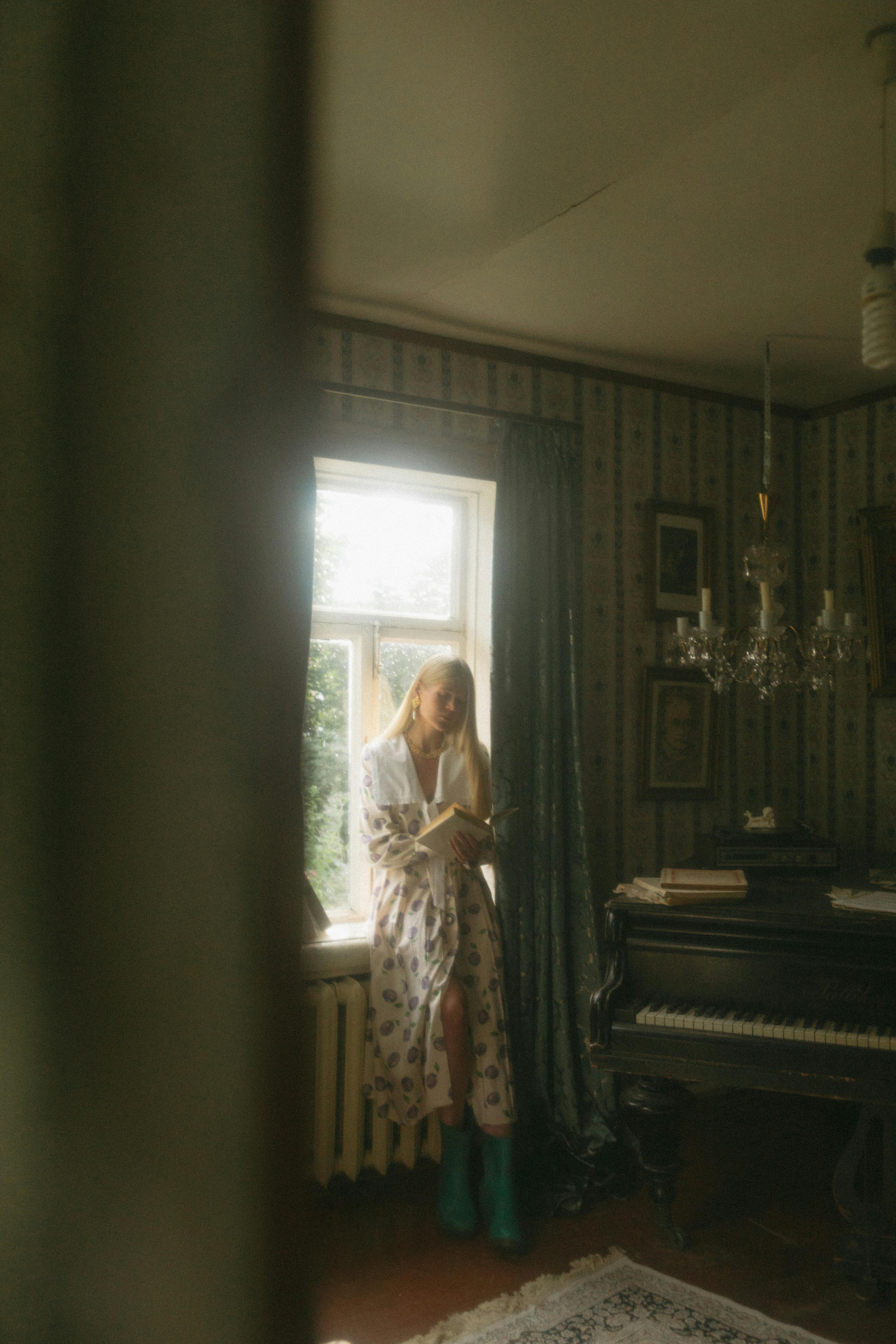 Woman in vintage room reading by window, wearing white dress.