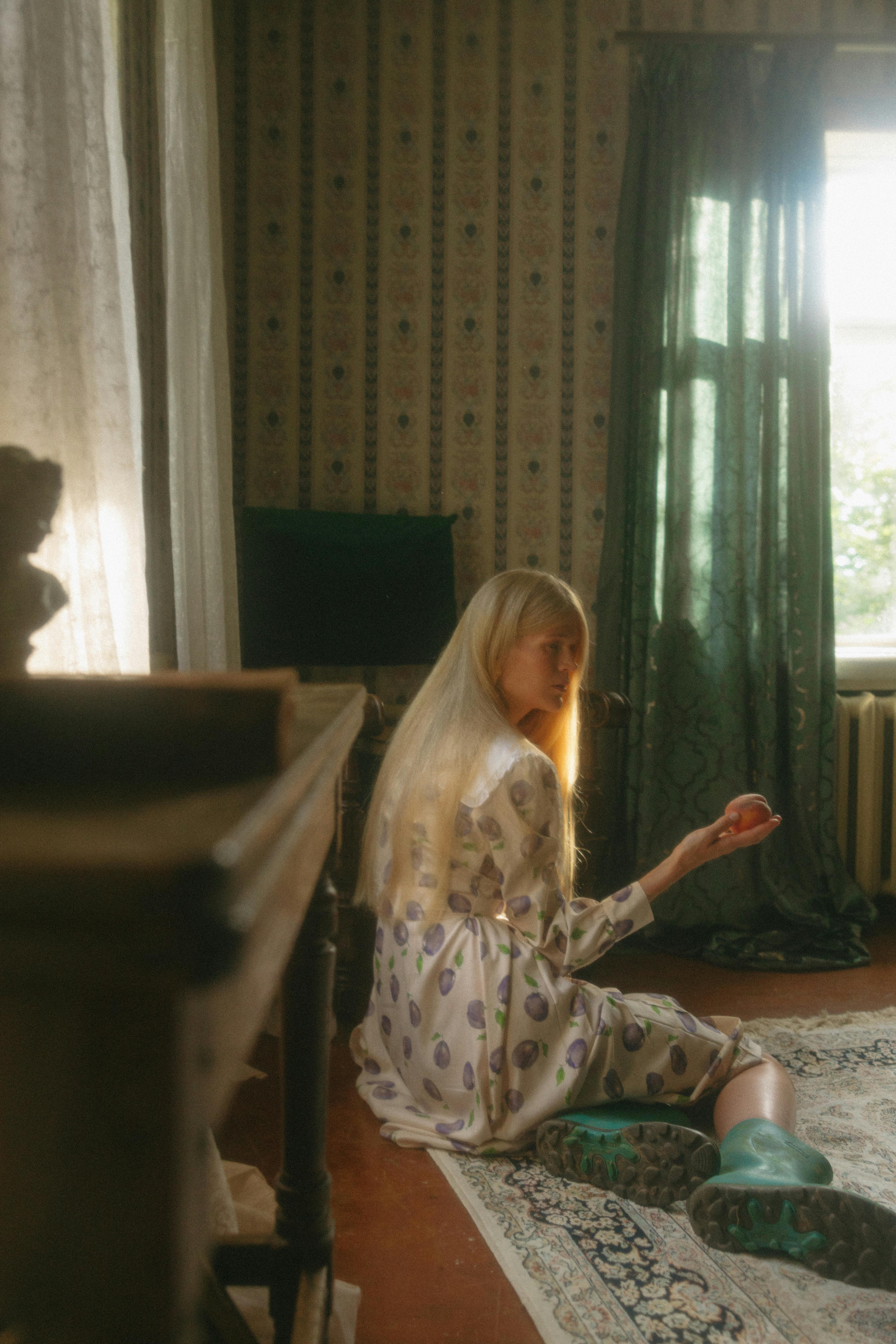 A woman with long blonde hair sits on a Persian carpet holding an apple in a vintage room.