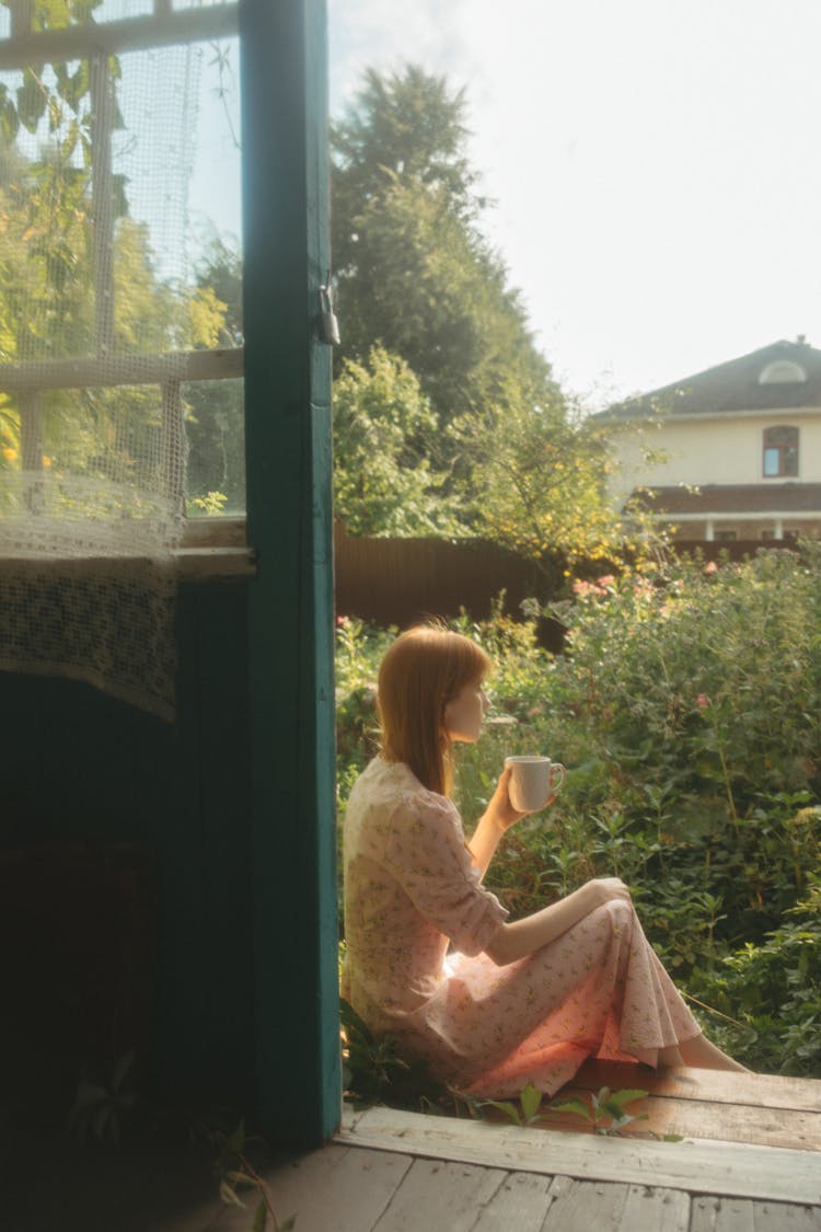 Woman In Pink Dress Sitting Outside The Door Near Green Plants