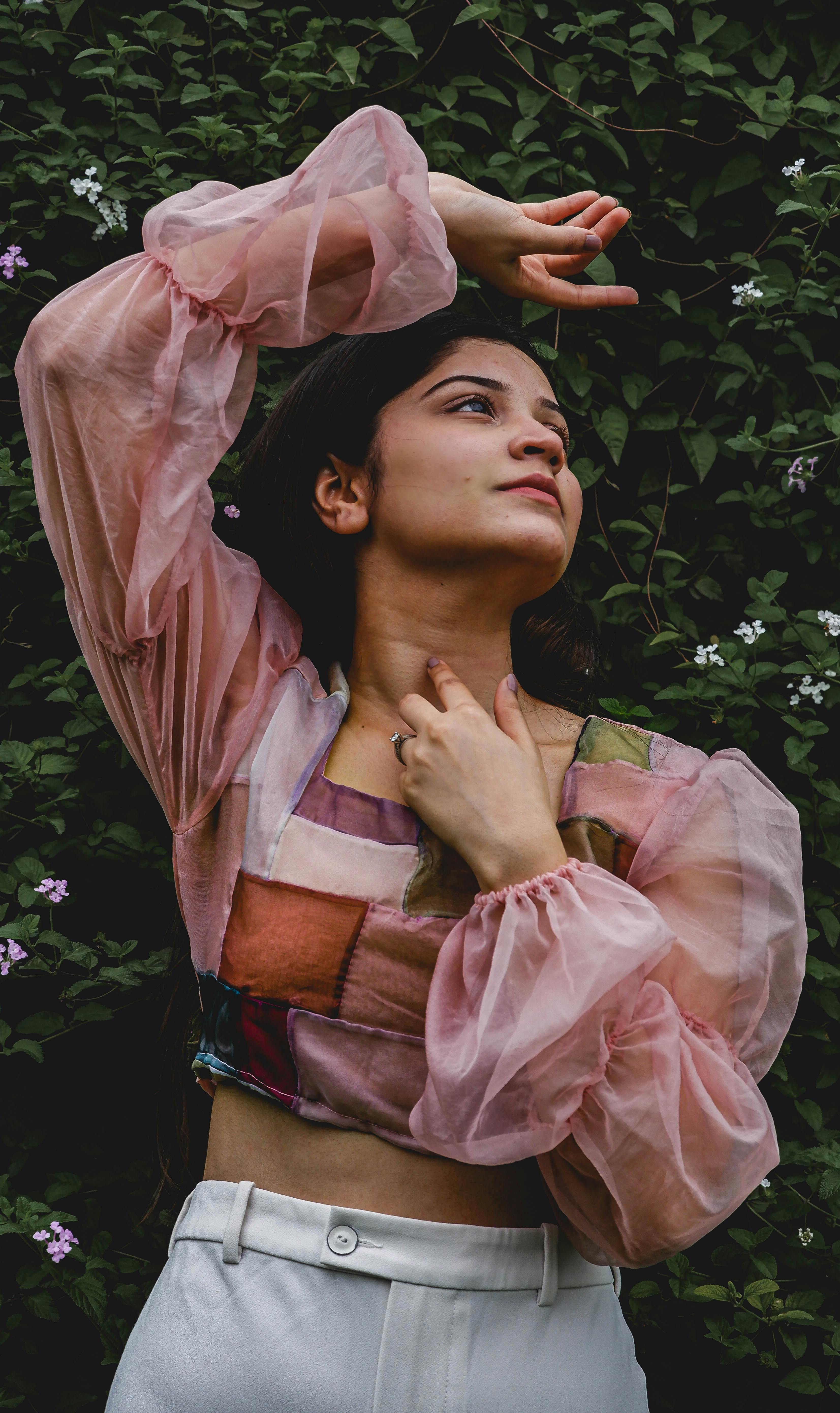A beautiful Asian woman in a pink chiffon crop top poses gracefully amidst flowering plants.