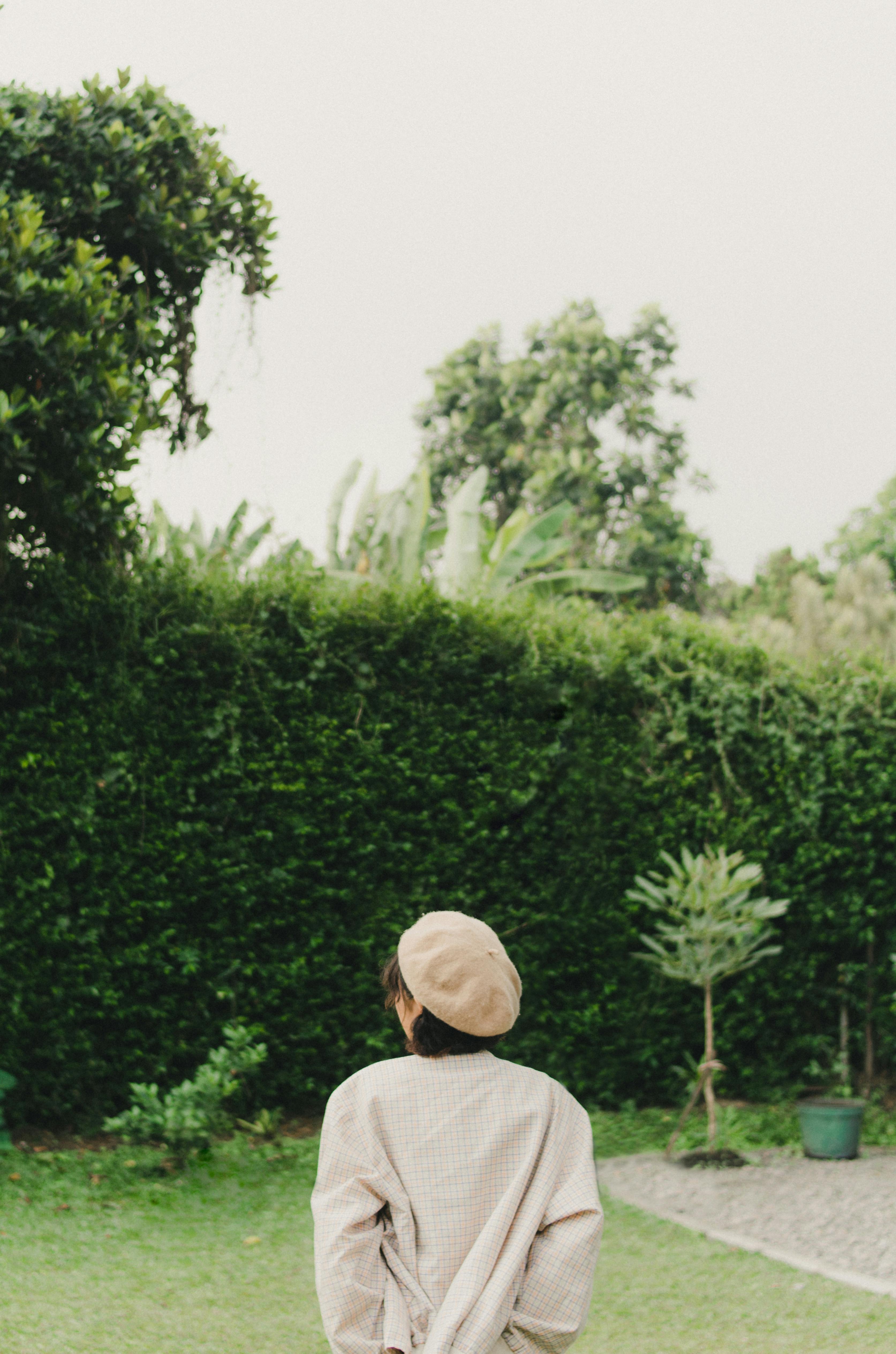 A woman wearing a beret stands alone, gazing at a lush green garden, conveying solitude and reflection.