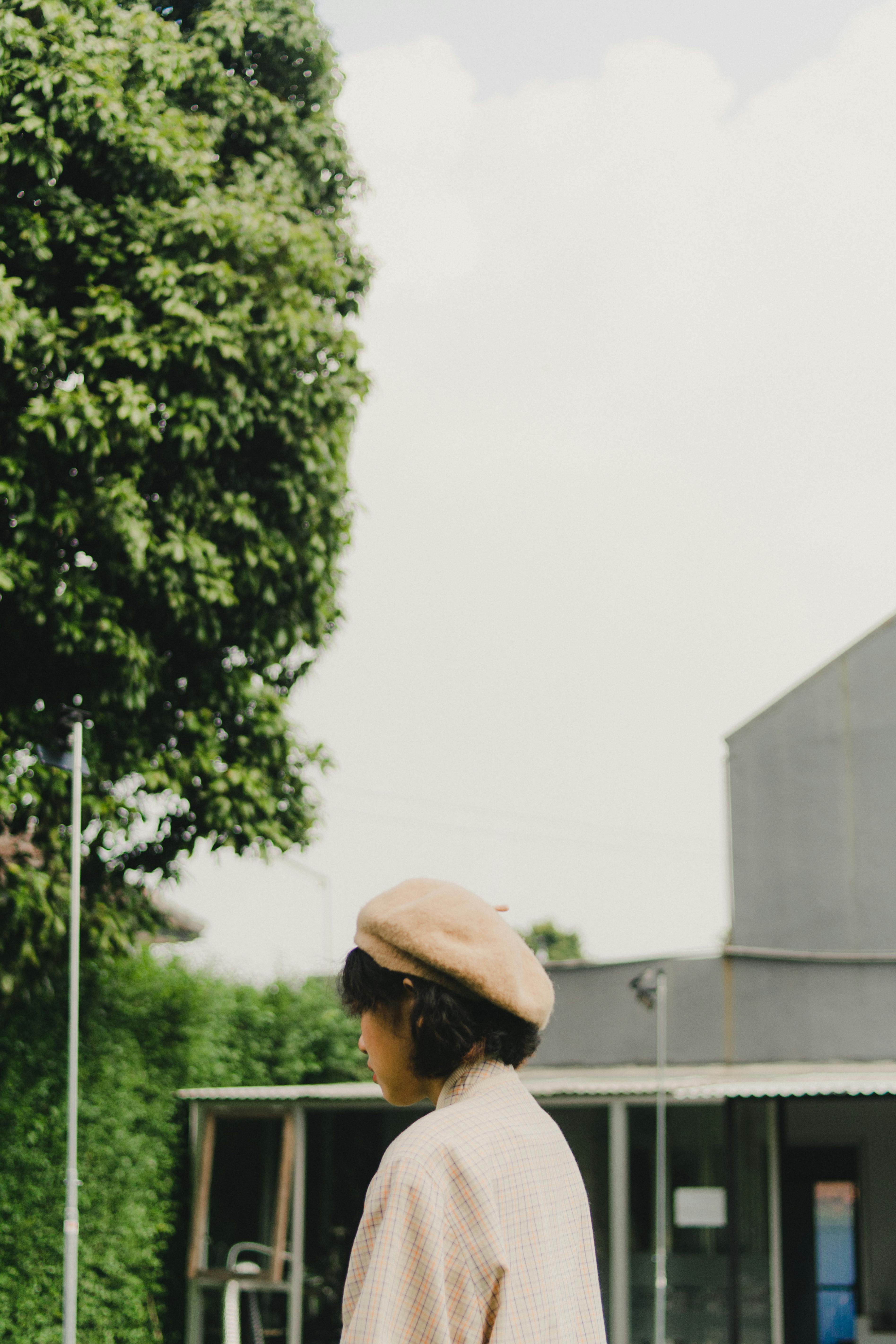 A person wearing a beret stands outdoors near a building under a bright sky.