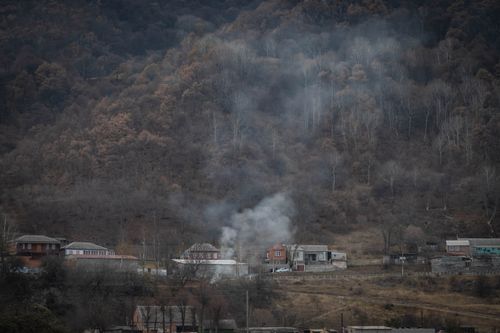 Free A tranquil autumn village scene with smoke rising amidst dense forest-covered mountains, creating a mysterious atmosphere. Stock Photo