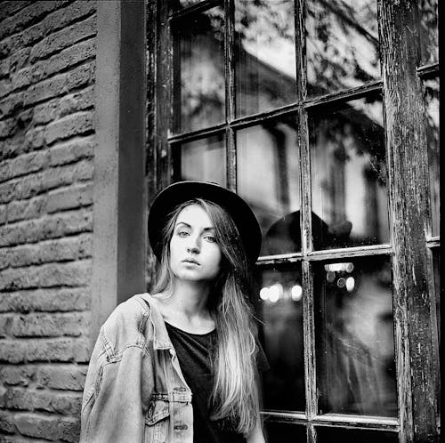 무료 창에 기대어 여성의 데님 재킷과 모자의 회색조 사진 스톡 사진