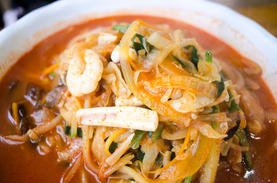 A steaming bowl of red-broth jjampong noodle soup with mussels, shrimp, squid rings, and wheat noodles, topped with sliced scallions and a red chili sheen on the surface.