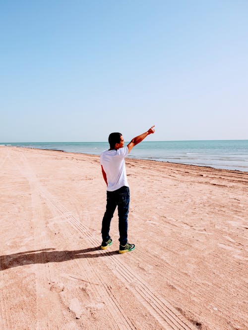 A Man in White Shirt Standing on the Beach Sand while Pointing His Finger in the Sky