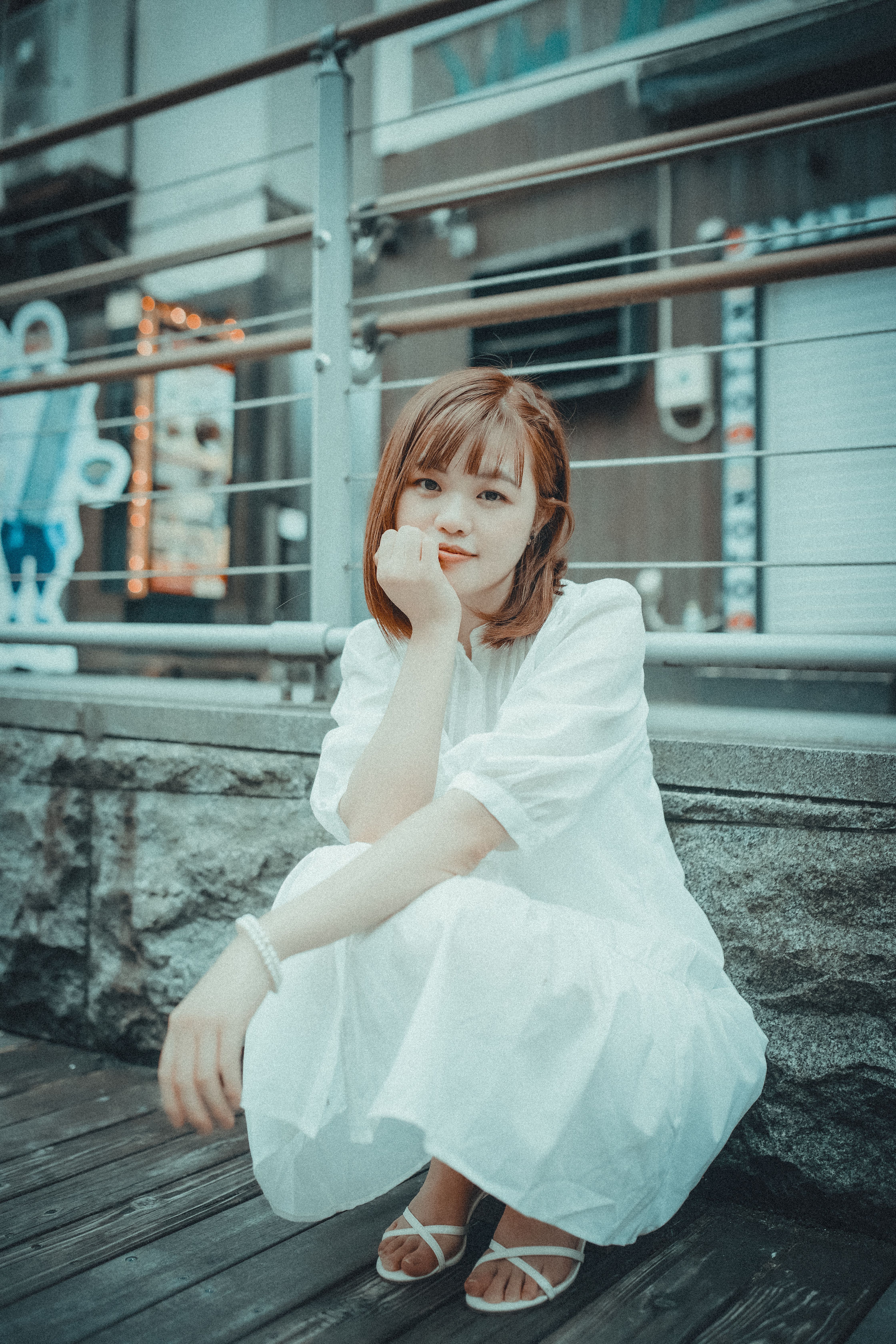 A serene adult woman in a white dress sits outdoors, smiling gently in an urban setting.